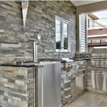 Outdoor stainless-steel kitchen with built-in grill, fridge, sink, and stone veneer counter and wall panels.