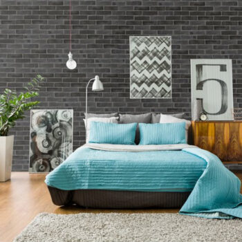 Modern bedroom with a turquoise quilted bedspread against a dark brick wall, three decorative pillows, and a shaggy rug in front of a wooden dresser with framed art pieces.