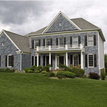 Front view of a two-story stone house with white columns, a balcony, and a green lawn.