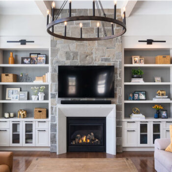 Living room with a stone fireplace, a mounted flat-screen TV, circular chandelier, and built-in white shelves.