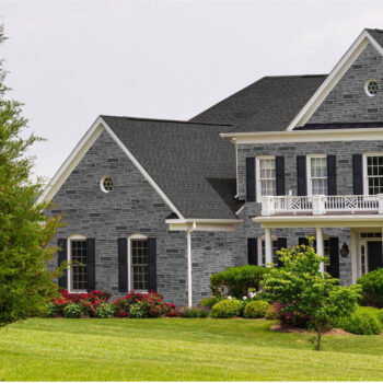 Gray stone two-story house with black shutters, white balcony railing, and a neatly landscaped front yard with flower beds.