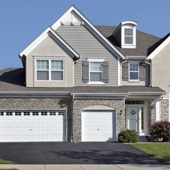 Front view of a suburban two-car garage home with stone veneer, gray siding, and white doors, including a decorative front entrance and window shutters under a clear blue sky.