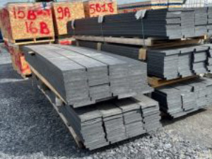 Stacks of metal planks on pallets at an outdoor storage yard with orange tags in the background.